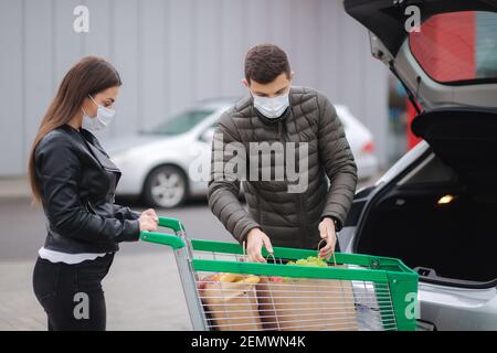 Giovane coppia in maschere di carico borse in tronco dopo supermercato shopping durante la quarantena. Uomo con sua moglie nel parcheggio all'aperto Foto Stock
