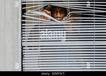 Giovane uomo che guarda fuori dalle persiane a casa Foto Stock