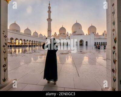 Una giovane donna musulmana che indossa un abaya nero prende un selfie in una bellissima moschea al tramonto ad Abu Dhabi, Emirati Arabi Uniti. Foto Stock