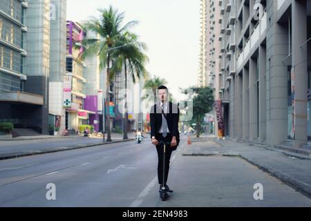 Uomo d'affari asiatico che guida uno scooter elettrico sulle strade della città per andare a lavorare al mattino. Spostamenti quotidiani che riflettono meglio il mondo del lavoro di to Foto Stock