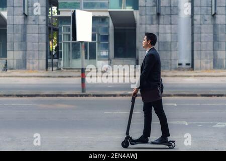 Uomo d'affari asiatico che guida uno scooter elettrico sulle strade della città per andare a lavorare al mattino. Spostamenti quotidiani che riflettono meglio il mondo del lavoro di to Foto Stock