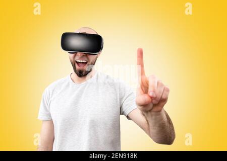 Occhiali VR. Un uomo bearded meravigliato in occhiali di realtà virtuale, con la sua bocca leggermente aperta, punta un dito nello spazio. Sfondo giallo. Il concep Foto Stock