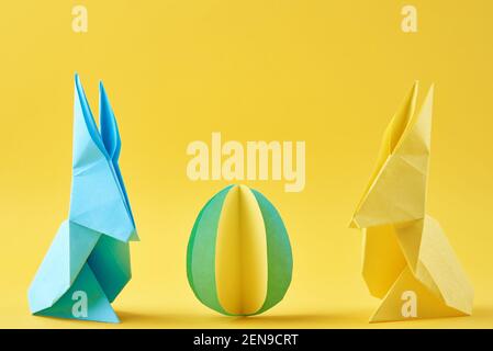 Due conigli di carta origami Esater e uovo colorato su sfondo giallo. Concetto di celebrazione pasquale Foto Stock