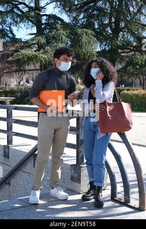 Giovane coppia latina che indossa una maschera protettiva che guarda e parla sullo smartphone. Nuova normalità nel campus universitario. Foto Stock