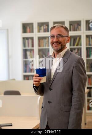 Uomo d'affari felice di media età in piedi alla scrivania dell'ufficio durante la pausa caffè. Ritratto di uomo maturo con tazza di caffè. Foto Stock