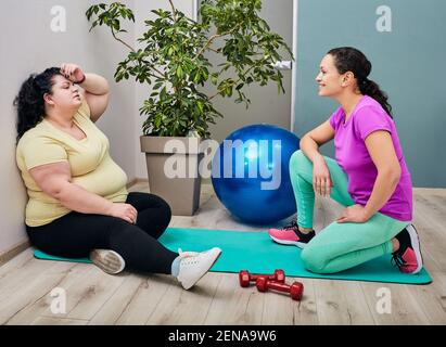 Donna stanca in sovrappeso seduta in palestra dopo l'allenamento con il suo allenatore Foto Stock