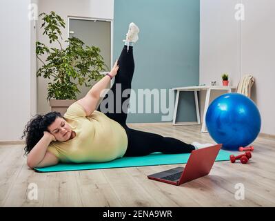 Allenamento di stretching fitness e pilates online. Donna sovrappeso che fa un allenamento per le gambe che si trovano sul tappetino fitness durante la visione di tutorial online al lapt Foto Stock