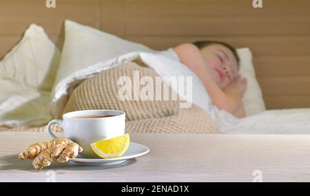 Una tazza di tè su un piatto con radice di zenzero e una fetta di limone su sfondo sfocato. Dormire bambino malato in letto. Foto Stock