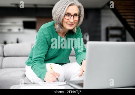 Vivace donna invecchiata grigio capelli occupato come un'ape, elegantemente vestito, seduto al computer portatile, loft in stile appartamenti, scrivere sulla carta, tenendo la penna in mano e digitando sulla tastiera, lavorando duramente Foto Stock