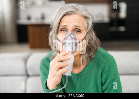 Donna anziana dai capelli grigi, ha molte rughe, si sente male, tenendo la maschera di ossigeno, mancanza di ossigeno, sensibile all'aria inquinata, seduta a casa su un auto-isolamento, guarda falcamente, facendo inalazione Foto Stock