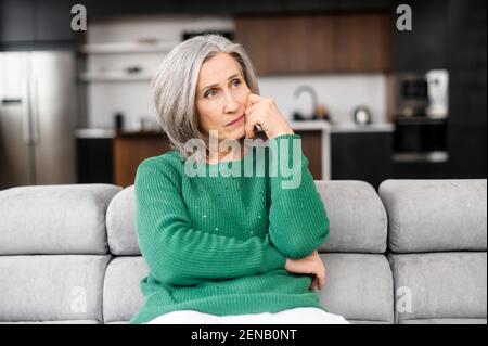 Donna anziana premurosa con capelli grigi, pensando al futuro, progettando la vita, preoccupata per i suoi figli, casa e proprietà, toccando il viso, guardando da parte, seduto sul divano puro in appartamento Foto Stock