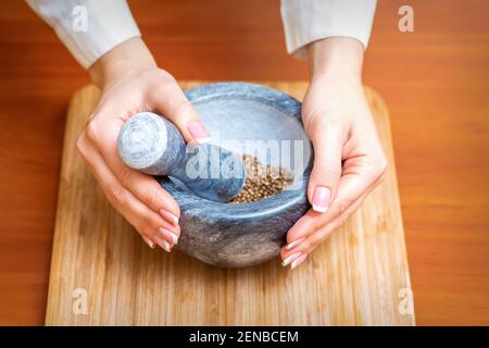 Le mani femminili macinano le spezie secche del pepe in mortaio su a. tavola da cucina in legno Foto Stock