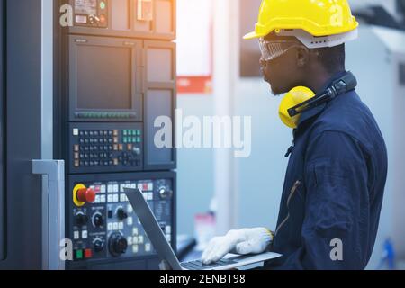 Uomo di lavoro con casco giallo e tastiera per la digitazione con protezione dell'orecchio di computer portatile in fabbrica Foto Stock