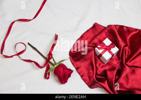 Rosa rossa con nastro rosso e tessuto rosso satinato con scatola gif su letto bianco con cuscino. San Valentino o vacanza romantica background Foto Stock