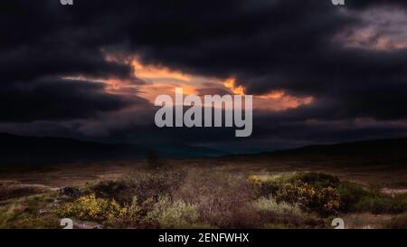 Condizioni atmosferiche e di illuminazione estreme durante il tramonto nelle Highlands scozzesi. Cielo scuro e suggestivo con nuvole nere e una finestra di luce solare arancione. Foto Stock
