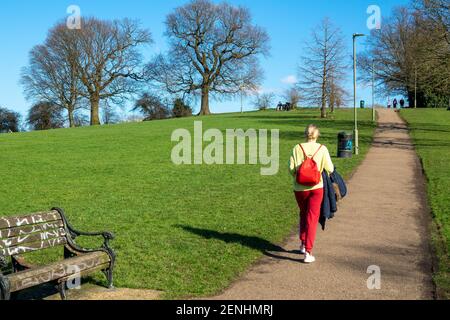 Donna bionda che indossa pantaloni rossi e jumper giallo cammina su una collina in Sunny Hill Park, Mill Hill / Hendon, godendo del sole in una giornata invernale cielo blu e soleggiata. Inizio di un weekend soleggiato a Londra. Regno Unito Meteo. Londra, Regno Unito. 26 febbraio 2021. Foto Stock