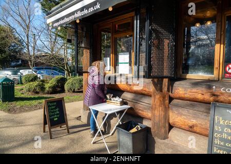 Donna che ordina un drink e un cibo in un locale all'aperto. Code socialmente distanziate di persone che ordinano bevande al Sunny Hill Cafe, una caffetteria a Sunny Hill Park, Hendon. Inizio di un weekend soleggiato con cielo blu.. Regno Unito Meteo. Londra, Regno Unito. 26 febbraio 2021. Foto Stock