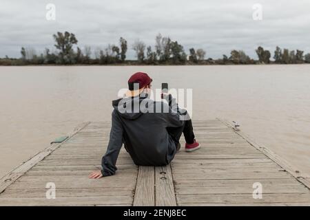 Giovane uomo da dietro indossando un maglione grigio e un berretto rosso utilizzando uno smartphone e scattando foto un molo vicino ad un fiume in una giornata nuvolosa Foto Stock