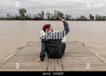 Giovane uomo da dietro indossando un cappuccio rosso e un il pullover grigio è seduto in un molo accanto a a. fiume che prende selfie in una giornata nuvolosa Foto Stock