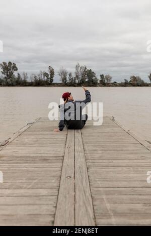 Giovane uomo da dietro indossando un cappuccio rosso e un il pullover grigio è seduto in un molo accanto a a. fiume che prende selfie in una giornata nuvolosa Foto Stock