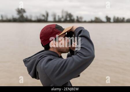 Giovane uomo che indossa un pullover grigio e berretto rosso è scattare foto in un molo vicino ad un fiume Foto Stock