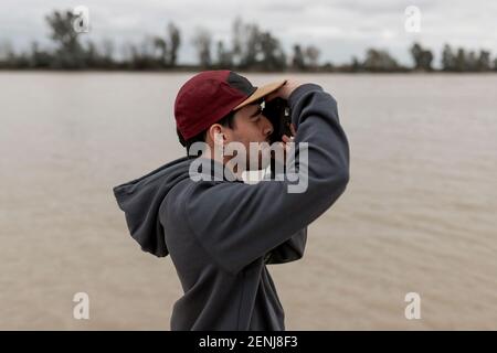 Giovane uomo che indossa un pullover grigio e berretto rosso è scattare foto in un molo vicino ad un fiume Foto Stock