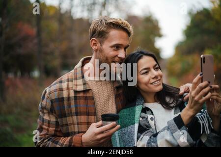 Felice coppia bere tè e scattare foto sul cellulare mentre fare un picnic nella foresta d'autunno Foto Stock