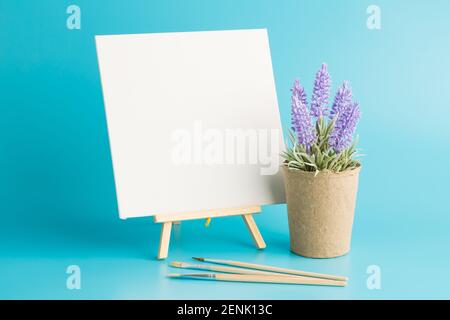 Cavalletto in legno con tela bianca, spazzole e fiori di lavanda su sfondo blu. Foto Stock