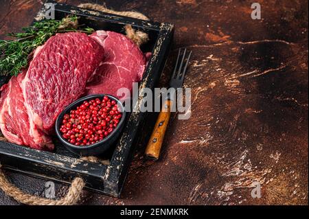 Bistecca di manzo fresco di Chuck Raw Eye roll in un vassoio di legno con erbe. Sfondo scuro. Vista dall'alto. Spazio di copia Foto Stock