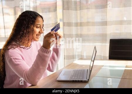 Felice giovane ragazza che acquista online con la sua carta di credito e il computer a un tavolo vicino alla finestra. Foto Stock