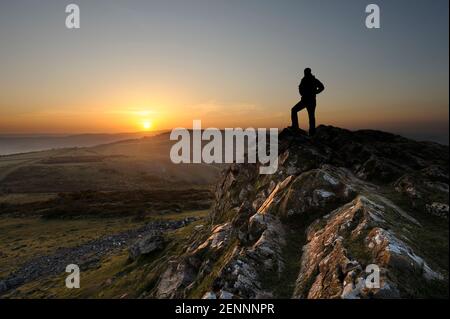 Un uomo solista guarda il sole che sorge dalla cima della cresta rocciosa sul picco di Crook sulle colline di Mendip a Somerset, Regno Unito. [La persona nell'immagine è Foto Stock