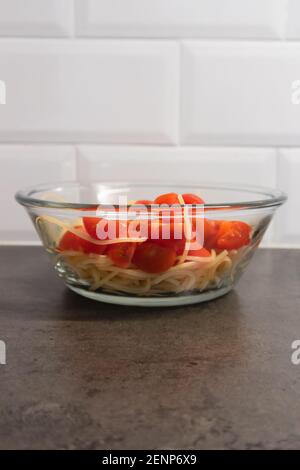 Ciotola in vetro piena di spaghetti e pomodori ciliegini tagliati, verticale. Piastrelle bianche sullo sfondo Foto Stock