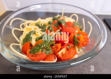 Ciotola di vetro piena di spaghetti e pomodori ciliegini tagliati con pesto di basilico verde sulla parte superiore. Idee per il pranzo vegano Foto Stock