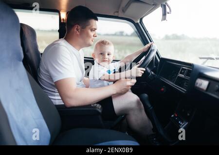 Padre che guida una macchina con il suo figliolo. Ragazzino seduto sulle gambe del padre e girando il volante in macchina. Felice padre e figlio Foto Stock