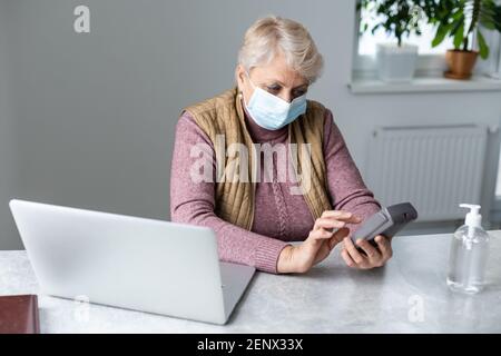 donna anziana seduta con computer portatile e terminale pos durante la quarantena Foto Stock