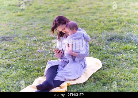 Bella giovane donna in abito grigio seduta sull'erba che tiene il suo bambino di sei mesi mentre controlla i social media smartphone. Concept stile di vita Moth Foto Stock