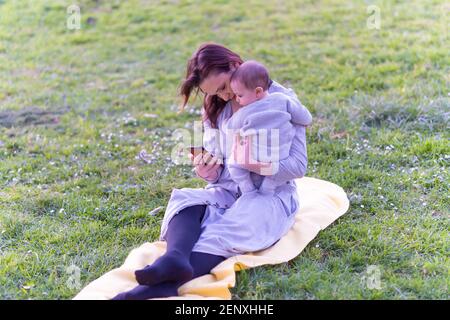 Bella giovane donna in abito grigio seduta sull'erba che tiene il suo bambino di sei mesi mentre controlla i social media smartphone. Concept stile di vita Moth Foto Stock