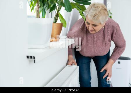 Donna anziana che cerca di mantenere caldo riscaldando le mani il radiatore di riscaldamento in inverno Foto Stock