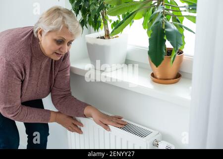 Donna anziana che cerca di mantenere caldo riscaldando le mani il radiatore di riscaldamento in inverno Foto Stock