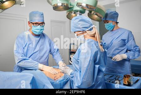 Gruppo di medici in uniformi chirurgiche blu che fanno chirurgia estetica in sala operatoria. Squadra chirurgica che esegue chirurgia estetica e utilizza strumenti medici in clinica. Concetto di chirurgia plastica. Foto Stock