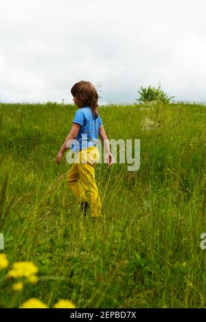 Un ragazzo di sei anni in pantaloni gialli e una T-shirt blu con lunghi capelli cammina nel prato. Verde erba e cielo. Foto Stock