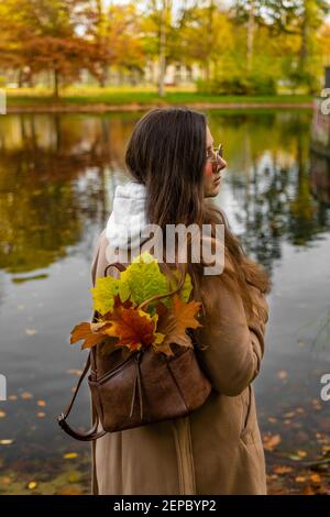 Giovane donna dai capelli scuri in cappotto marrone in piedi presso lo stagno dopo ha raccolto le foglie colorate asciutte nel parco autunnale Foto Stock