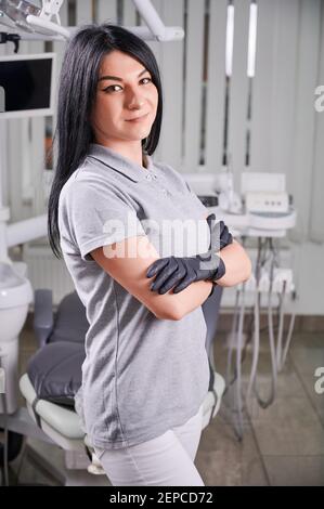 Ritratto di dentista femminile in piedi in un ufficio ben attrezzato, sorridente, indossando uniforme e guanti neri. Concetto di assistenza sanitaria e ortodontica Foto Stock