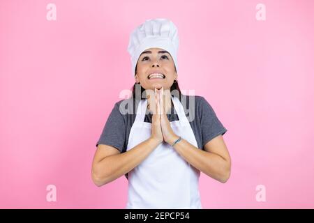 Giovane donna ispanica che indossa uniforme da panettiere su sfondo rosa che elemosina e pregare con le mani insieme con l'espressione della speranza sul volto molto emozionale an Foto Stock