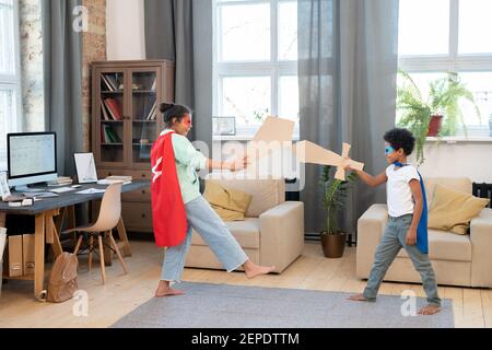 Due adorabili fratelli in costumi di supereroi che si divertono il pavimento mentre si gioca con spade di cartone al centro di soggiorno Foto Stock