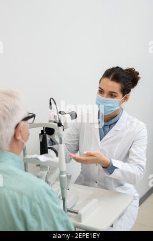 Giovane oftalmologo femminile in bianco e maschera protettiva seduta accanto apparecchiature mediche mentre andare a testare la vista di un maschio anziano paziente Foto Stock