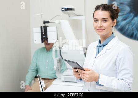 Felice giovane oftalmologo femminile in whitecoat che ti guarda mentre uso della compressa sullo sfondo del paziente con la sua vista controllata su Foto Stock