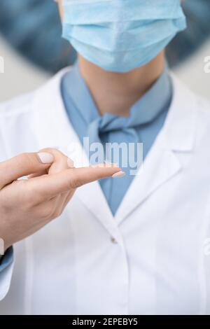 Mano di giovane oftalmologo femminile in bianco e maschera protettiva tenendo la lente di contatto sulla punta dell'indice davanti a. fotocamera Foto Stock