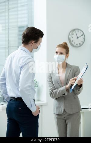 Due giovani eleganti ragionieri in maschere protettive che discutono di documento finanziario con diagrammi e grafici alla riunione di lavoro in ufficio Foto Stock