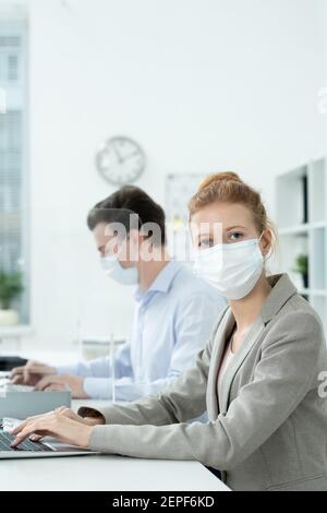 Giovane donna direttore d'ufficio in maschera protettiva ed elegante grigio vestito guardando voi mentre la rete sullo sfondo di maschio collega Foto Stock
